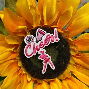 May include: An embroidered patch featuring the word Cheer! in pink script lettering, accompanied by a small pink megaphone icon, a pom-pom, and a stylized silhouette of a cheerleader in a skirt, all outlined in white thread.