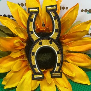 May include: An embroidered patch featuring two interlocking horseshoe shapes in yellow and black thread. The design includes white accent stitching along the edges and small black square details arranged symmetrically on the horseshoe surfaces.
