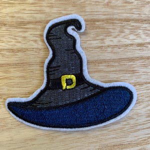 May include: Embroidered patch of a witch's hat. The hat is primarily dark gray with a blue brim and a white outline. A gold-colored buckle is on the hat's band. The patch is likely for clothing or accessories.