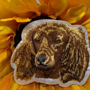 May include: Embroidered patch featuring a detailed brown and gold dachshund dog face. The patch is set against a bright yellow sunflower, creating a striking contrast. The dog's fur is intricately stitched, showcasing its features.