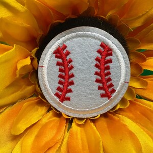 May include: A baseball-themed patch with red stitching on a gray background, set against a vibrant yellow sunflower. The baseball patch is round and features the classic red stitching design.