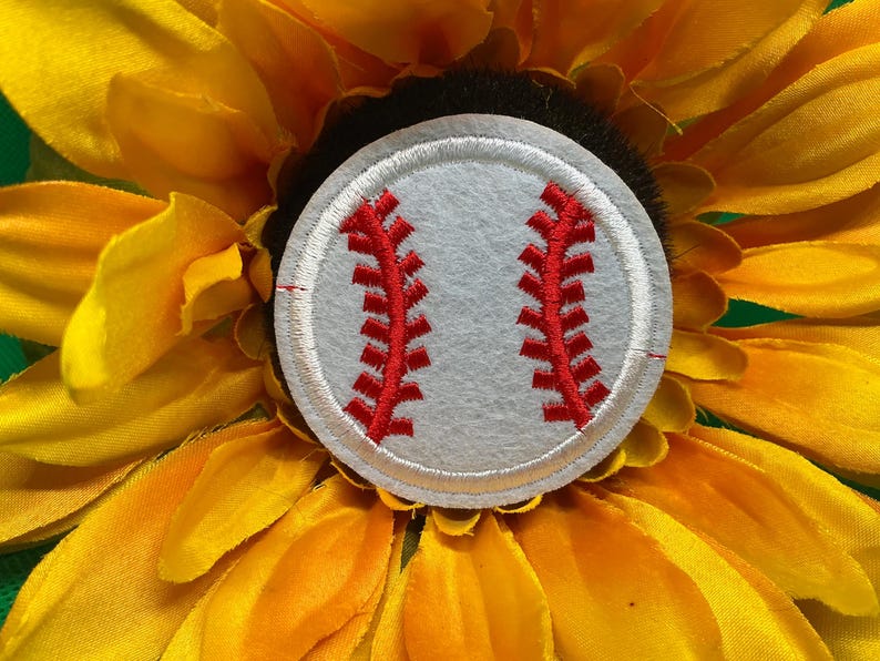 May include: A baseball-themed patch with red stitching on a gray background, set against a vibrant yellow sunflower. The baseball patch is round and features the classic red stitching design.