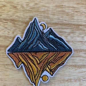 May include: Embroidered patch featuring a mountain range design. The upper half depicts blue mountains with a crescent moon, while the lower half shows a golden, textured landscape. The patch has a white border.