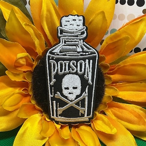 May include: An embroidered patch shaped like a bottle with a cork stopper. The patch is black with white stitching, featuring the word POISON in capital letters above a white skull and crossbones graphic. The design is outlined in white thread.