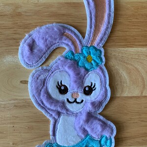 May include: A purple plush bunny with a blue flower on its head and a blue tutu. The bunny has a white face with black eyes and a pink nose.