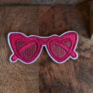 May include: A pink embroidered patch in the shape of heart-shaped sunglasses. The patch has a white border and a textured appearance. The lenses have a mesh-like design. The patch is on a wooden surface.