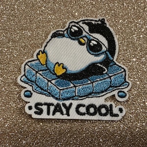 May include: Embroidered patch featuring a cartoon penguin wearing sunglasses, lounging on a blue ice block. The penguin is black, white, and yellow, with the text "STAY COOL" in white below. The patch has a white border.