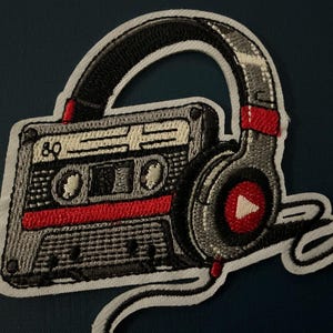 May include: Embroidered patch featuring a retro cassette tape and headphones design. The cassette is gray with red accents and the number "80". The headphones are gray with red details and a play button symbol. The patch has a white border.