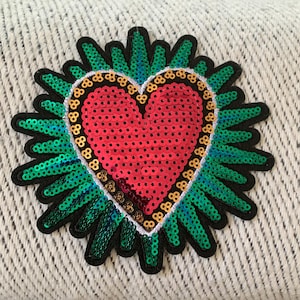 May include: A red heart-shaped sequin patch with a green and blue sequin border. The patch is surrounded by a black outline and has a white stitching detail.