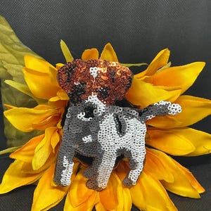 May include: A sequined dog figurine in black, white, and brown, positioned on a bright yellow sunflower. The dog has a spotted pattern and is set against a dark background. The sunflower petals are a vibrant yellow.