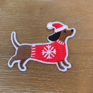 May include: Embroidered iron-on patch of a brown dachshund wearing a red sweater with a white snowflake design and a red Santa hat.