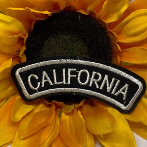 May include: A black and white embroidered patch with the word "CALIFORNIA" in white lettering. The patch is arched and set against a bright yellow sunflower.