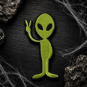May include: An embroidered patch featuring a lime green alien figure with a large oval head and black almond-shaped eyes. The alien is depicted in a standing position with one arm raised, displaying a peace sign gesture with two fingers extended.