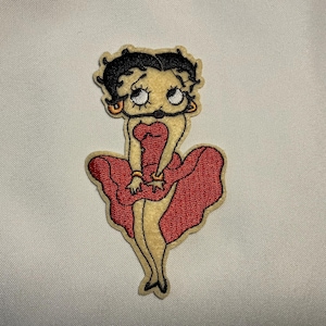 May include: Embroidered patch featuring a cartoon character in a red dress with a flared skirt. The character has black hair, large eyes, and is wearing gold hoop earrings. The patch is on a cream-colored background.