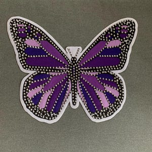 May include: Purple butterfly-shaped iron-on patch with rhinestones. The butterfly has a gradient of purple shades and is outlined in black.