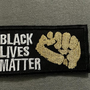 May include: Black rectangular patch with the text "BLACK LIVES MATTER" in white capital letters. A gold embroidered fist is on the right side of the patch. The patch is on a gray surface.