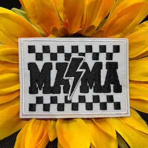 May include: A white patch with a black and white checkered pattern and the word "MAMA" in bold black letters with a lightning bolt in the center. The patch is set against a yellow sunflower.