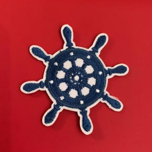 May include: Embroidered navy blue ship's wheel patch with white accents. The nautical-themed patch features a detailed design with eight spokes and a central circle. The patch is set against a vibrant red background.