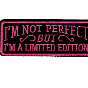 May include: A rectangular embroidered patch with a black background and a pink border. The text in pink embroidery reads I'M NOT PERFECT BUT I'M A LIMITED EDITION, featuring decorative flourishes between the lines of text on this fabric accessory.