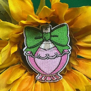 May include: Embroidered patch featuring a pink perfume bottle design with a green bow. The bottle has a heart-shaped body and is outlined in white. The patch is set against a yellow sunflower.