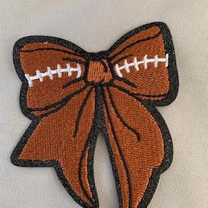 May include: A brown and black embroidered patch shaped like a bow with white stitching. The bow is made to look like a football.