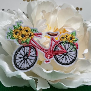 May include: An embroidered patch featuring a pink bicycle with black wheels. The bicycle has a front basket and a rear rack, both filled with yellow sunflowers and green leaves. The design includes a brown seat and red pedals on a white background.