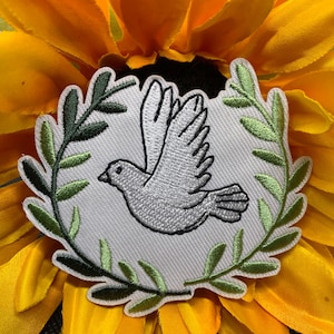 May include: Embroidered patch featuring a dove in flight, surrounded by a green olive branch wreath. The dove is gray, and the wreath is green. The patch is set against a bright yellow sunflower.