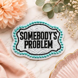 May include: An embroidered patch with a scalloped edge featuring the text SOMEBODY'S PROBLEM in bold black letters. The patch has a white background with a subtle swirling pattern, a black inner border, and a bright teal beaded-style outer trim.