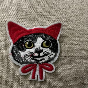 May include: An embroidered patch featuring a black and white cat face wearing a bright red hood. The cat has large, detailed green eyes, black whiskers, and a small black nose. A red bow is tied at the base of the cat's neck, completing the design.