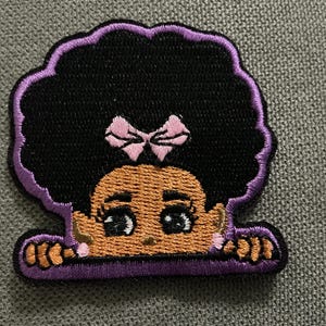 May include: Embroidered patch featuring a cartoon girl with black afro hair, a pink bow, and a tan face. The patch has a purple border and is designed to look like the girl is peeking over an edge. The patch is likely for clothing or accessories.