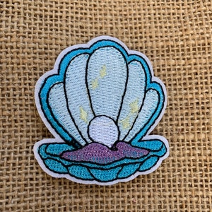 May include: A blue and white embroidered seashell patch with a pearl inside. The patch is on a burlap background.
