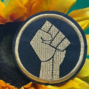 May include: Round, navy blue patch with a gold embroidered fist symbol, encircled by a gold border. The patch is set against a backdrop of yellow sunflower petals and a green background.