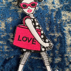 May include: Embroidered patch of a cartoon woman wearing pink sunglasses and a black and white polka dot dress. She is carrying a pink tote bag that says "LOVE" in white letters.