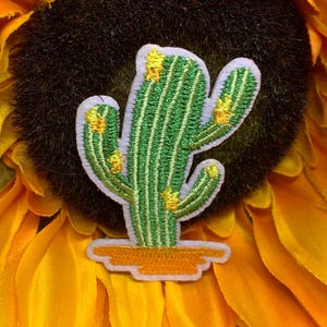 May include: Embroidered cactus patch with green and yellow details, set against a yellow and black background. The cactus has a white border and is placed on a yellow base. The patch is likely for clothing or accessories.