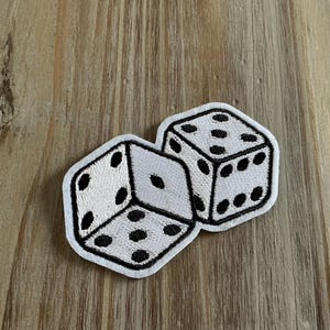 May include: Embroidered patch featuring two white dice with black dots. The dice are outlined in black and have a classic six-sided design. This patch is suitable for clothing, bags, or other fabric items.