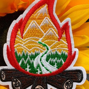 May include: Embroidered patch in the shape of a campfire flame. The patch features a mountain scene with a winding road, green trees, and a yellow and orange sky. The flame is outlined in red and white, with brown logs at the base.