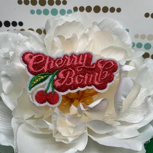 May include: An embroidered patch featuring the red text Cherry Bomb in a cursive font, accompanied by two red cherries with green stems and a small yellow leaf, all outlined in white thread, resting on a large white flower blossom.