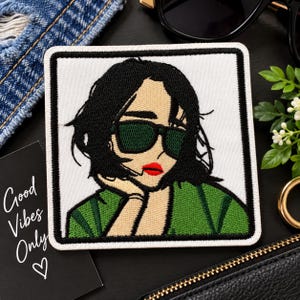 May include: A square embroidered patch featuring a stylized illustration of a person with short black hair, wearing dark green sunglasses and a green top. The person has bright red lips and is resting their chin on one hand. The design is outlined in black.