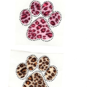 May include: Two paw print patches. One is pink with a leopard print design, outlined with silver rhinestones. The other is brown and tan with a leopard print design, also outlined with silver rhinestones. Both are on a white background.