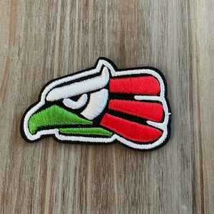 May include: Embroidered patch featuring a stylized eagle head in the colors of the Mexican flag: green, white, and red. The patch has a black border and is set against a wooden background.