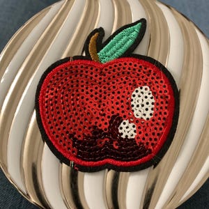 May include: A red apple patch with sequins, a green leaf, and a brown stem. The apple is on a white and gold striped background.