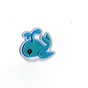 May include: A blue whale embroidered patch with a white outline and a black eye. The whale is facing left and has a small spout on top of its head.
