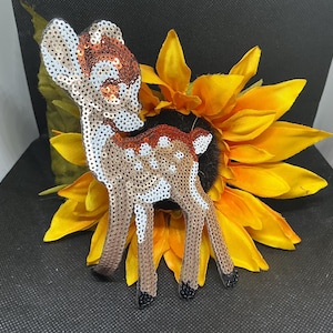 May include: A sequined deer figurine with brown, white, and copper-colored sequins, posed in front of a bright yellow sunflower. The deer has white spots and black hooves. The background is black.