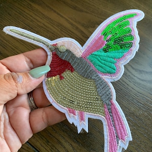 May include: A colorful sequined hummingbird patch with pink, green, gold, and gray accents. The patch is on a white background and has a white border.