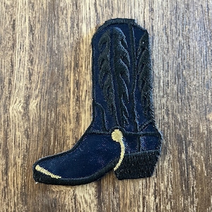 May include: A navy blue embroidered cowboy boot patch with gold accents. The boot features intricate stitching details and a classic western design. The patch is set against a wooden background.