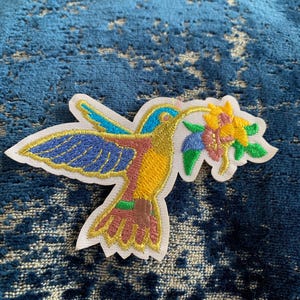 May include: A colorful embroidered patch of a hummingbird with a long beak, drinking nectar from a yellow flower. The hummingbird is blue, yellow, pink, and green. The flower is yellow and green.