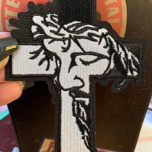 May include: Black and white embroidered patch in the shape of a cross, featuring a detailed depiction of Jesus wearing a crown of thorns. The patch has a hook and loop backing for easy attachment.