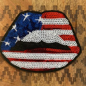 May include: A red, white, and blue sequined lip patch in the shape of a pair of lips. The patch is made of sequins and has a black felt backing.
