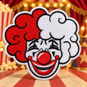 May include: An embroidered patch featuring a clown face with a split-color hairstyle, one side red and one side white. The clown has a bright red nose, a wide smiling mouth with black outlines, and closed eyes, all rendered in detailed thread work.