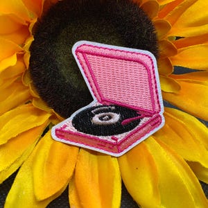 May include: A pink embroidered patch of a vintage record player with a black vinyl record. The patch has a white border and is set against a bright yellow sunflower.